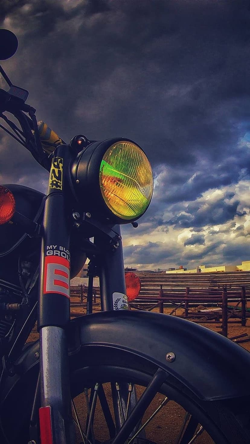 Vintage motorcycle headlight with yellow lens reflecting dark clouds, chrome forks, and black metal frame.