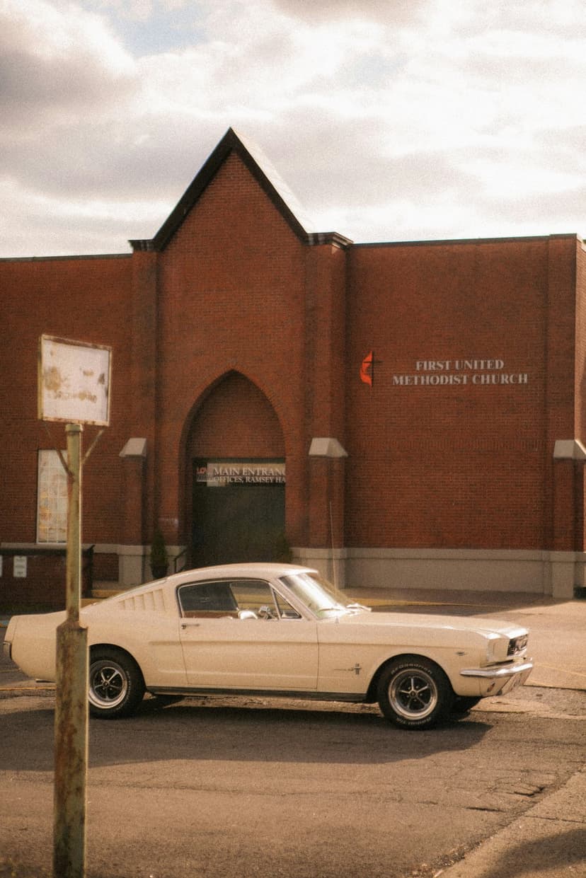 Cream Ford Mustang parked on a sunny street in front of a red brick Methodist church with an arched entryway.
