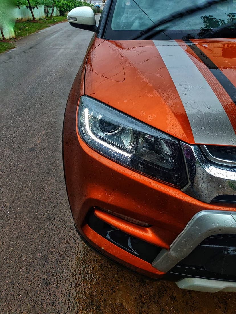 Orange sports car with white racing stripes covered in water droplets on a wet asphalt road during rainfall.