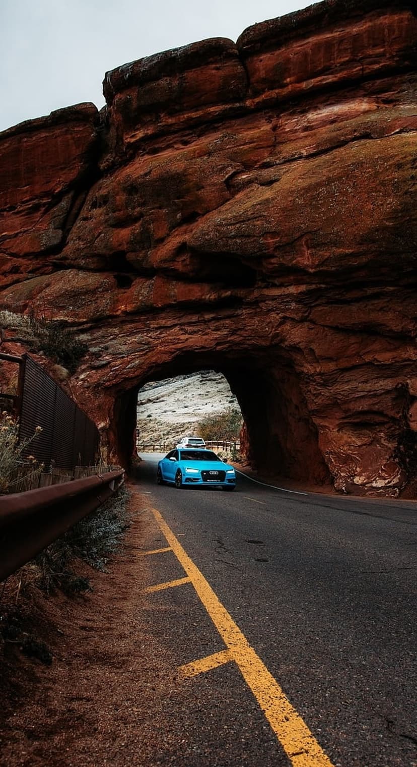 Blue Audi sedan driving through a narrow red sandstone rock tunnel on a paved road under an overcast sky.