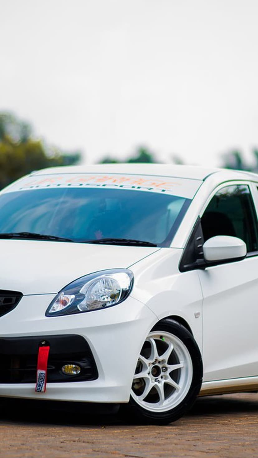 White Honda Brio hatchback with an orange sun visor, red tow hook, and multi-spoke wheels in an outdoor setting.