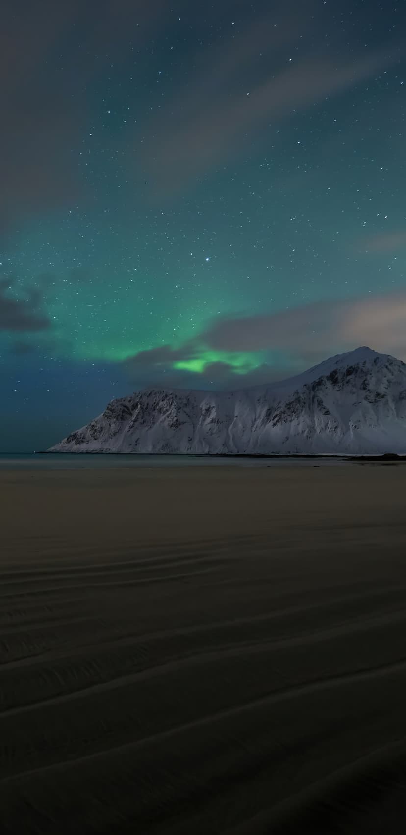 Emerald northern lights dancing over snow-capped mountains and rippled beach sand during a clear arctic night.