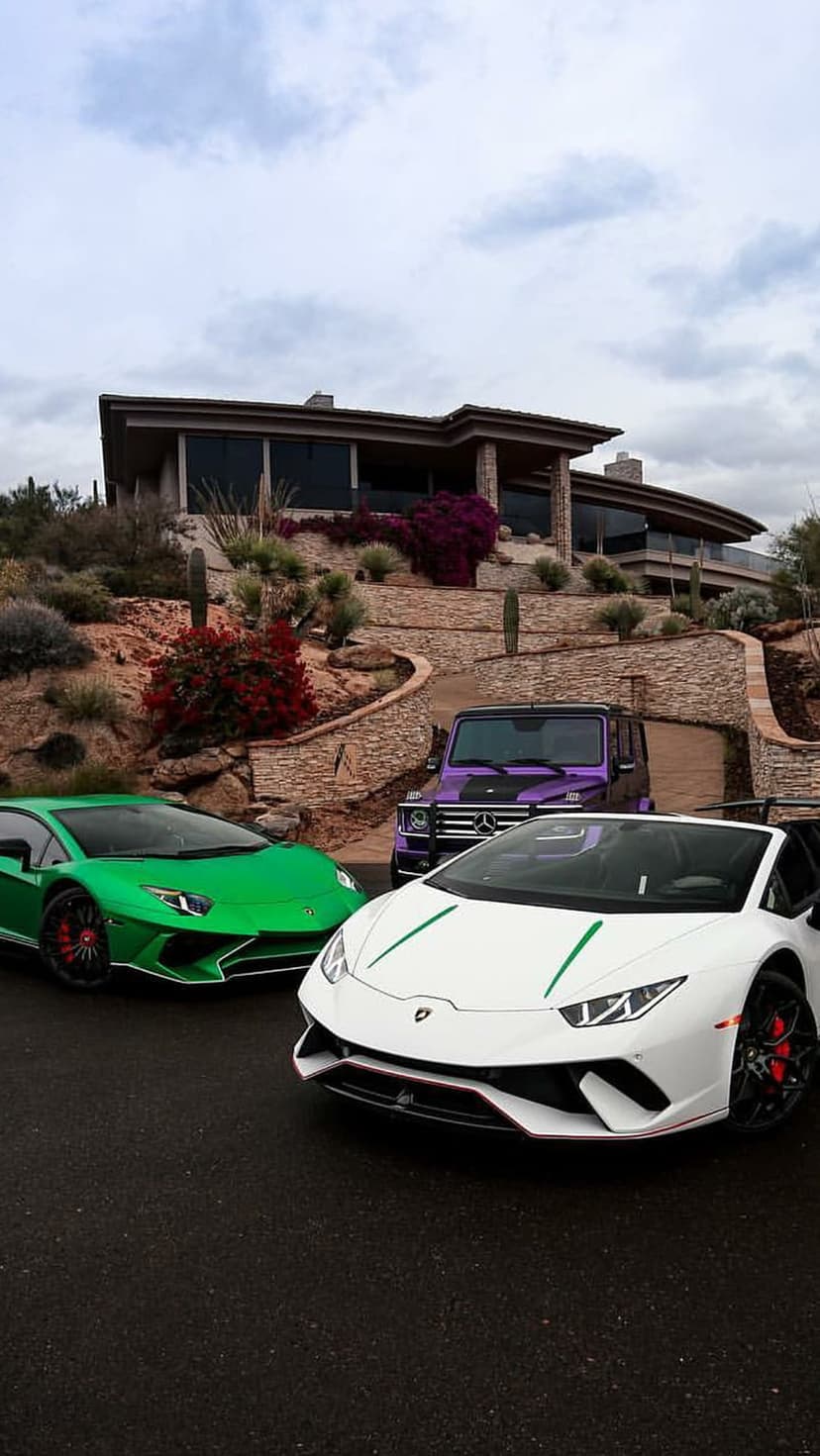 Green Lamborghini, white supercar, and purple G-Class parked at a modern desert home under a cloudy sky.