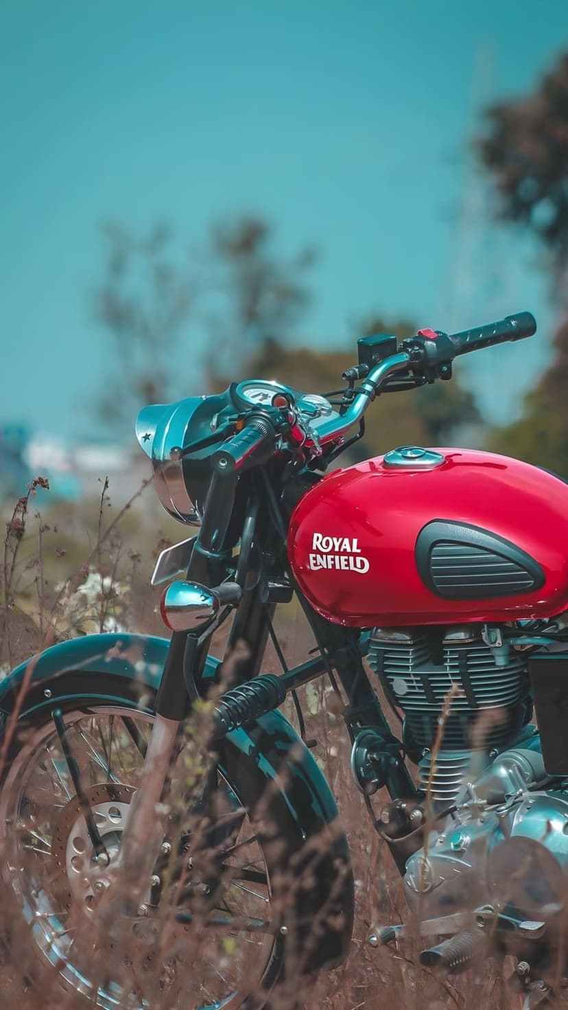 Red Royal Enfield motorcycle parked in tall dry grass with a chrome engine and blurred blue sky background.