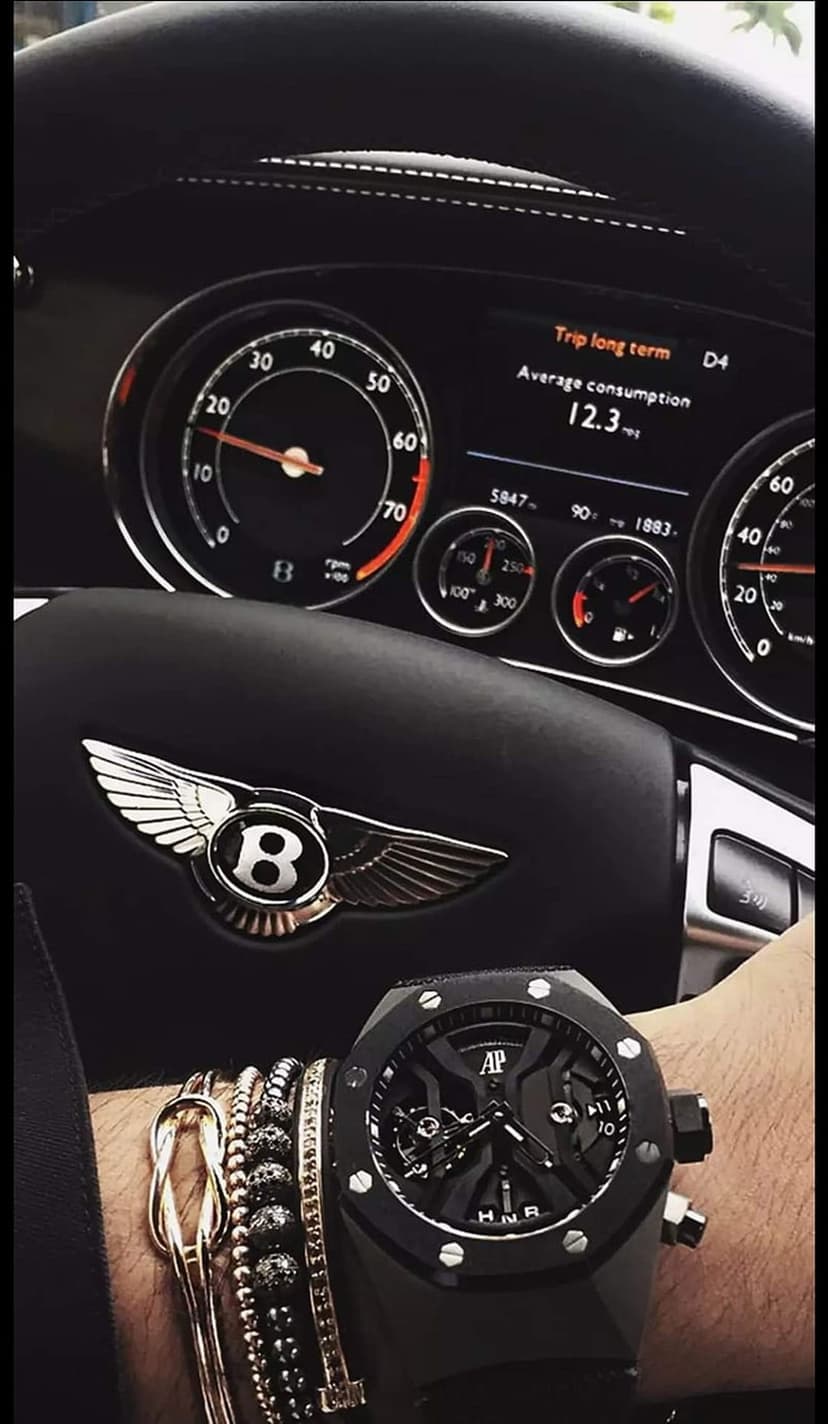 Bentley steering wheel with a silver winged emblem beside a luxury black watch on a wrist in a dim car interior.