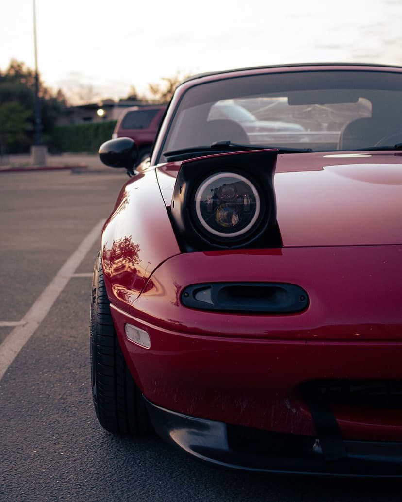 Red Mazda Miata with a raised pop-up headlight featuring an LED halo ring in a sunset parking lot.
