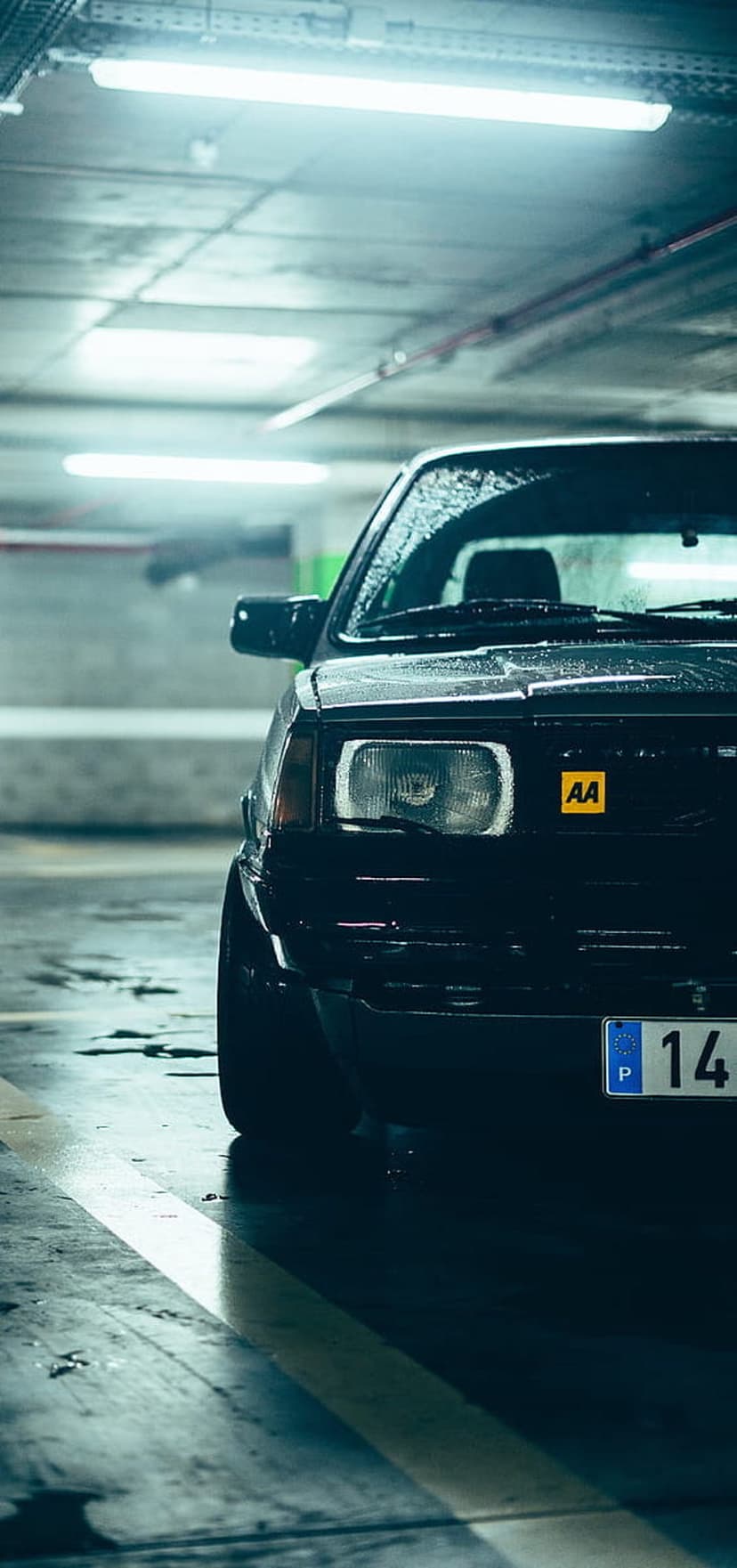 Wet black car with glowing headlights and water droplets parked in a dim blue lit parking garage at night.
