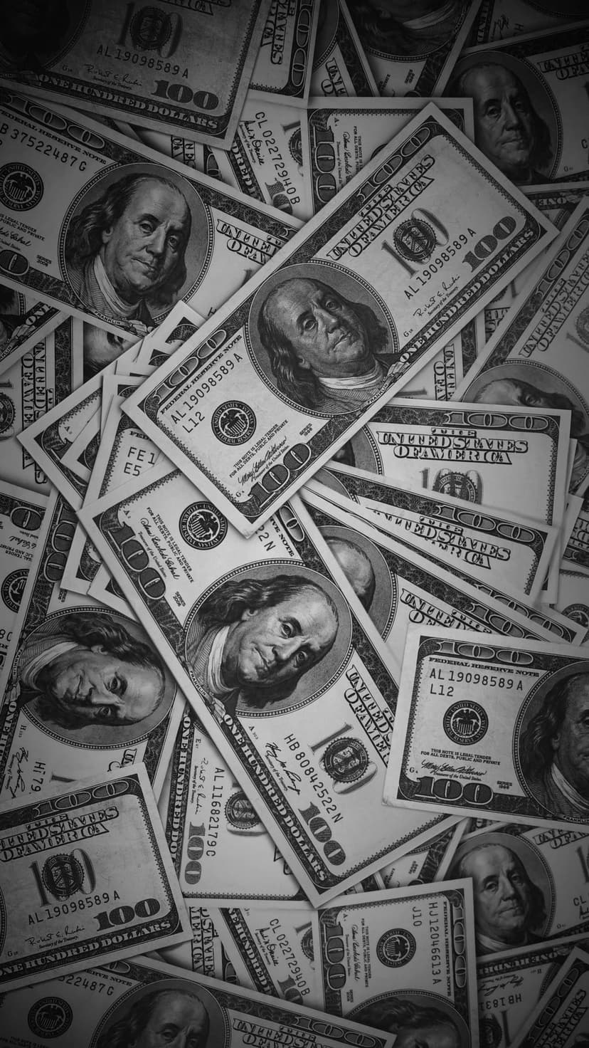 Overlapping US 100 dollar bills in a black and white overhead view featuring the portrait of Benjamin Franklin.