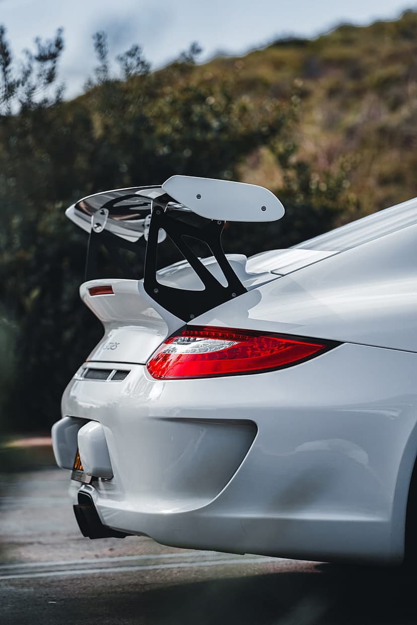 White Porsche 911 GT3 RS rear view features a swan-neck wing and glowing red taillights against blurred trees.