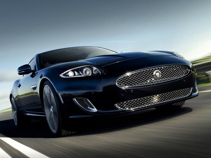 Dark blue Jaguar XK sports car driving on a road with blurred background and sparkling chrome grille.