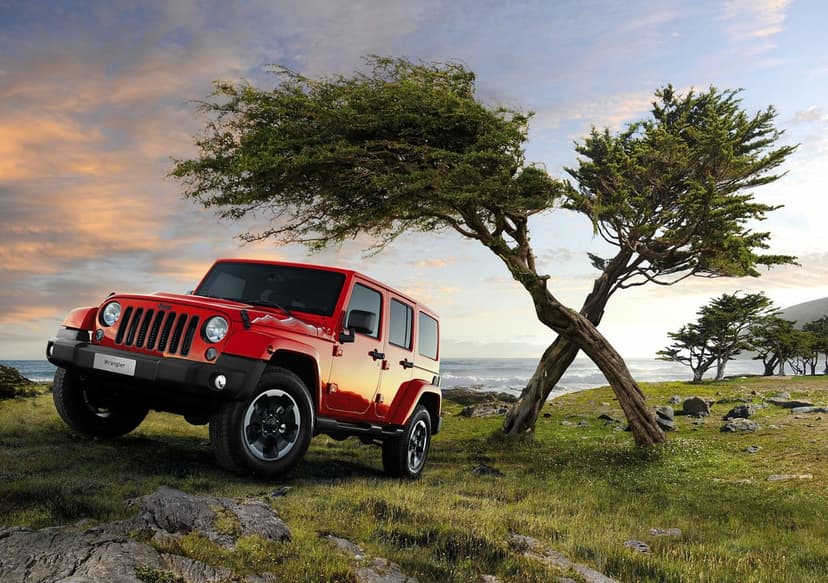 Red Jeep Wrangler parked on a grassy coastal cliff beside a wind-swept tree under a soft sunset sky.