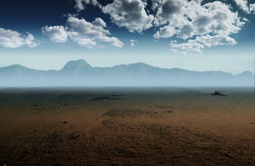 Arid desert valley features textured earth, a dark rock formation, and distant blue mountains under puffy clouds.