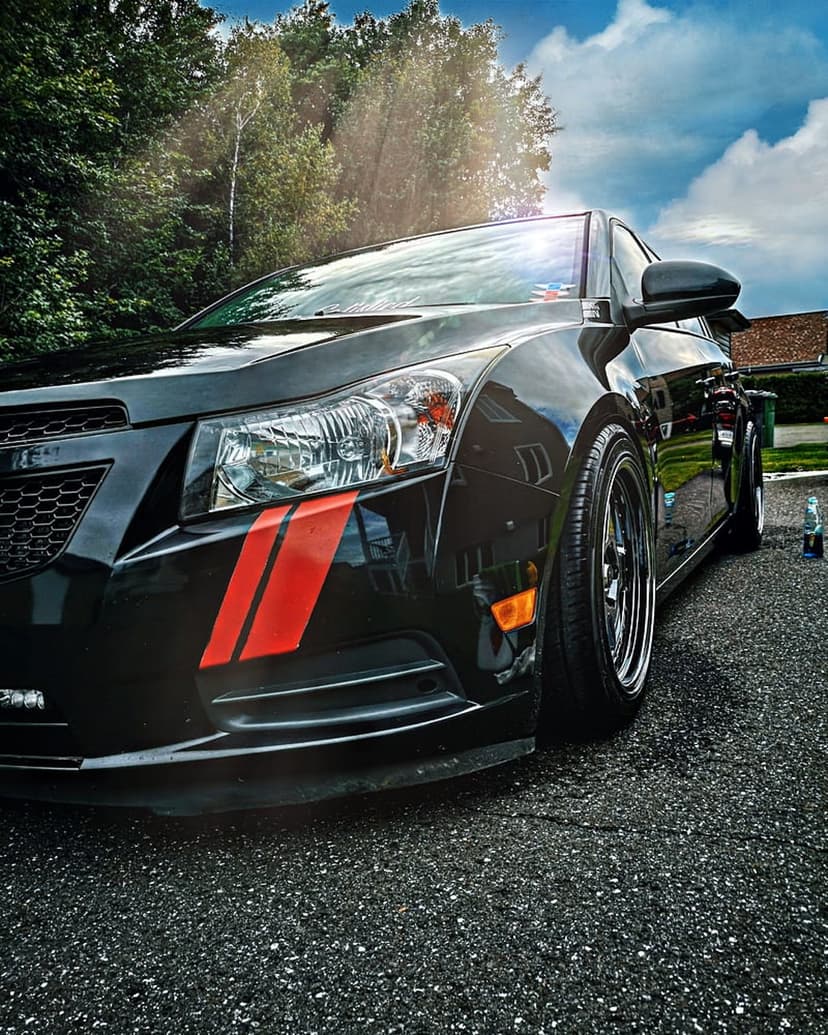 Black Chevrolet Cruze with red fender stripes sits on an asphalt driveway under a cloudy sky and green trees.