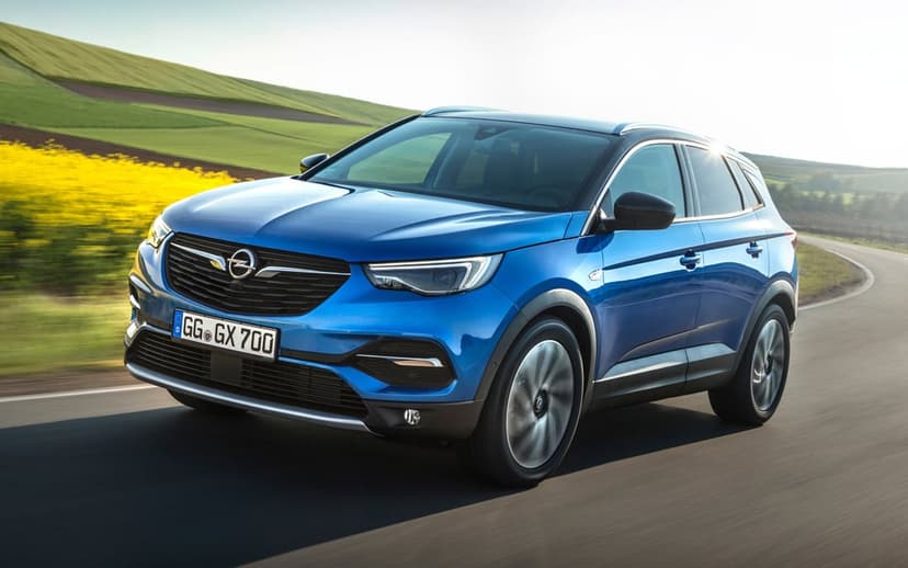 Blue Opel SUV driving fast on a winding road past green hills and yellow rapeseed fields under bright sunlight.