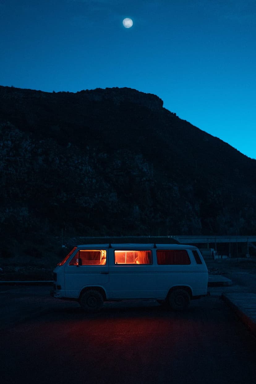Vintage white van with a glowing orange interior parked under a full moon and silhouetted mountain peaks.