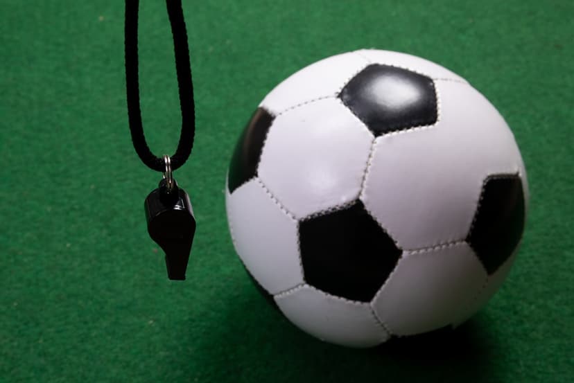 Soccer ball and a black whistle with a cord rest on a textured green surface under clear, bright lighting.