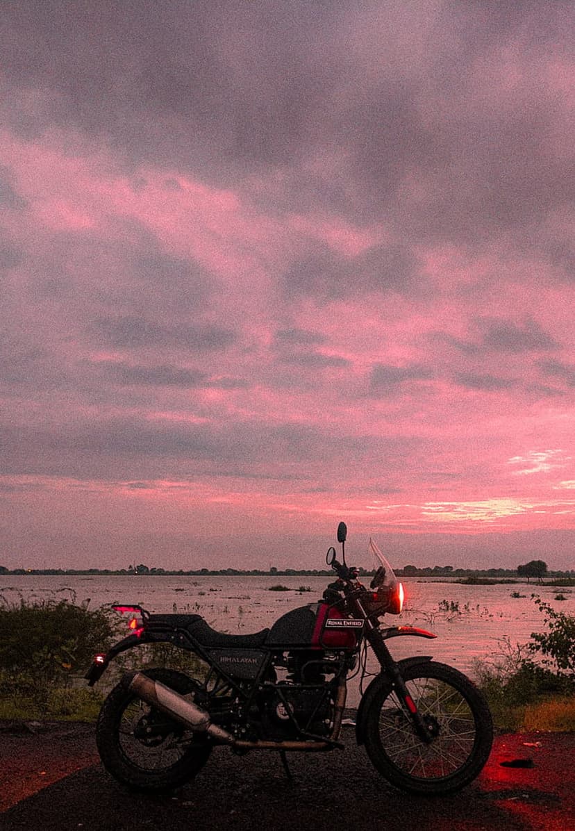 Royal Enfield Himalayan motorcycle parked on a lakeshore beneath a vibrant pink and purple twilight sky.