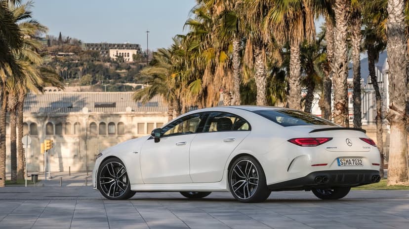 White Mercedes-AMG CLS 53 parked on a tiled plaza with palm trees and a historic hillside building background.