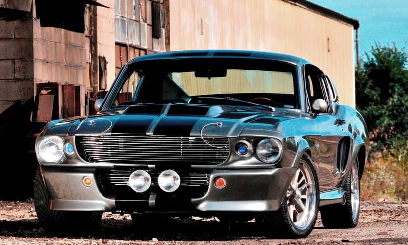 Gray 1967 Shelby GT500 Mustang with black racing stripes parked in front of a weathered industrial warehouse.