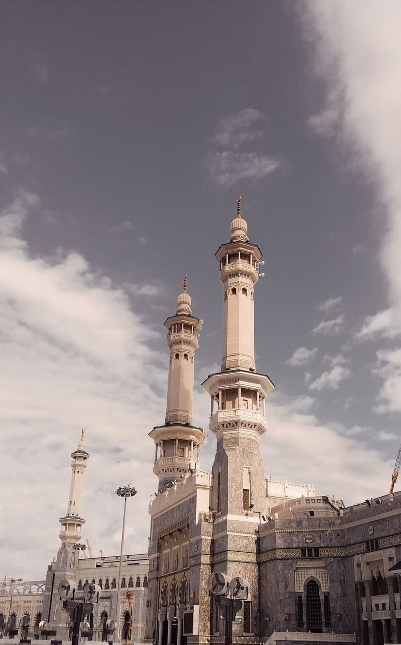 Stone minarets of Masjid Al-Haram stand against a backdrop of thick gray clouds under soft overcast lighting.