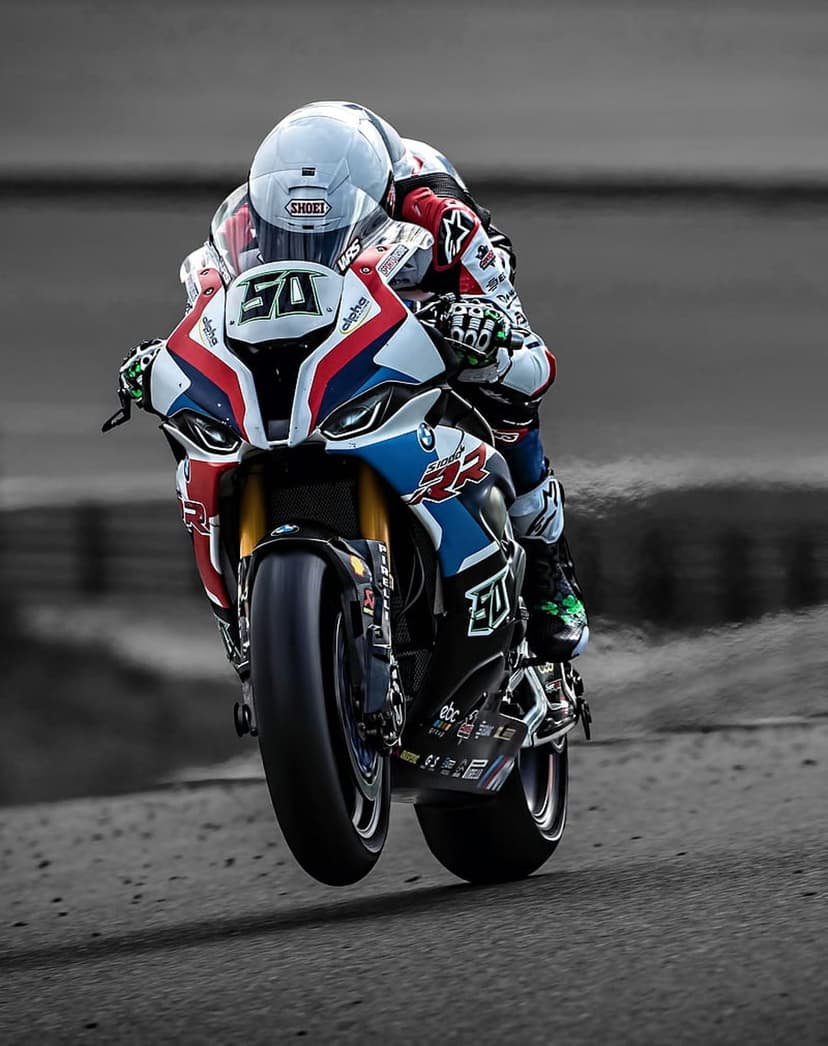 Racer in a tri-color suit performs a wheelie on a superbike with a white helmet on a blurred racing circuit.