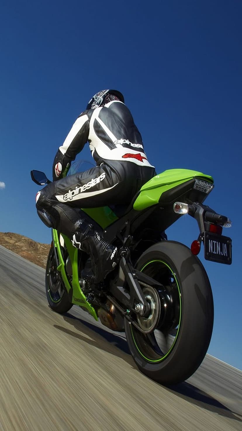 Motorcyclist in Alpinestars gear leans a green Kawasaki Ninja on a paved road under a bright blue sky.