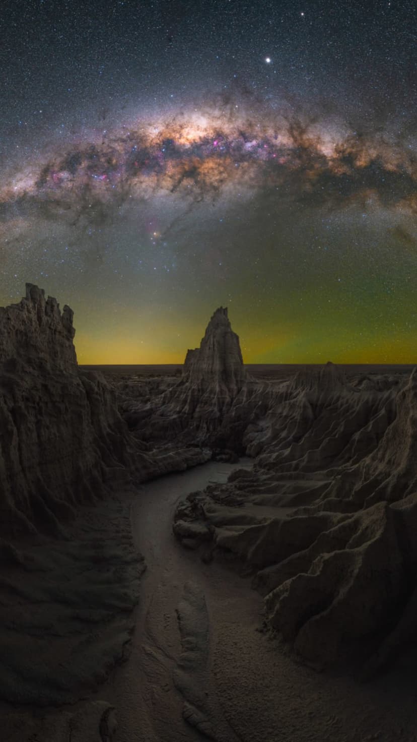 Milky Way galaxy arching over silhouetted desert rock formations and a winding trail under a starry night sky.