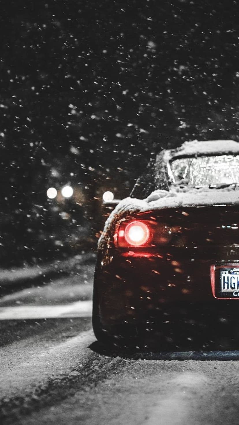 Red car tail light glowing through falling snowflakes on a dark winter night with soft bokeh background lights.
