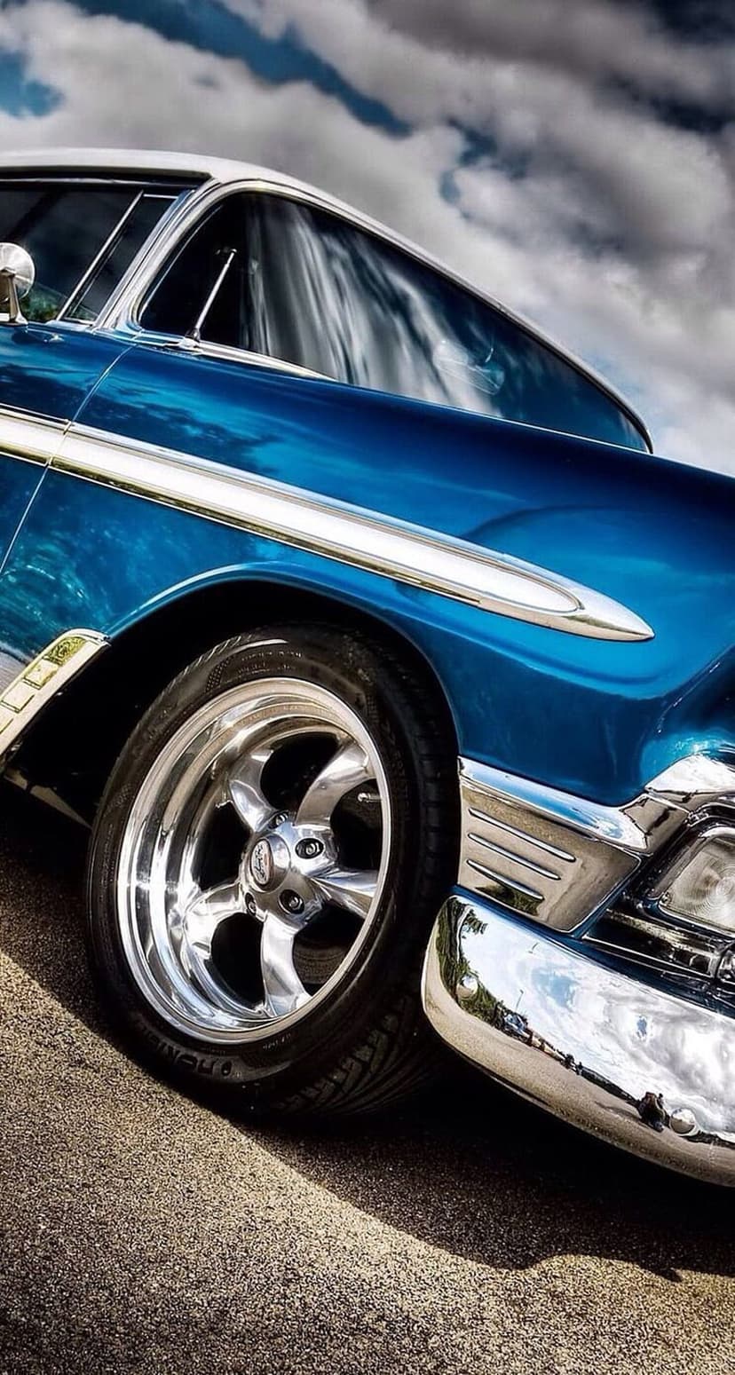 Blue vintage Chevrolet car features gleaming chrome wheels and polished metallic trim under a moody cloudy sky.