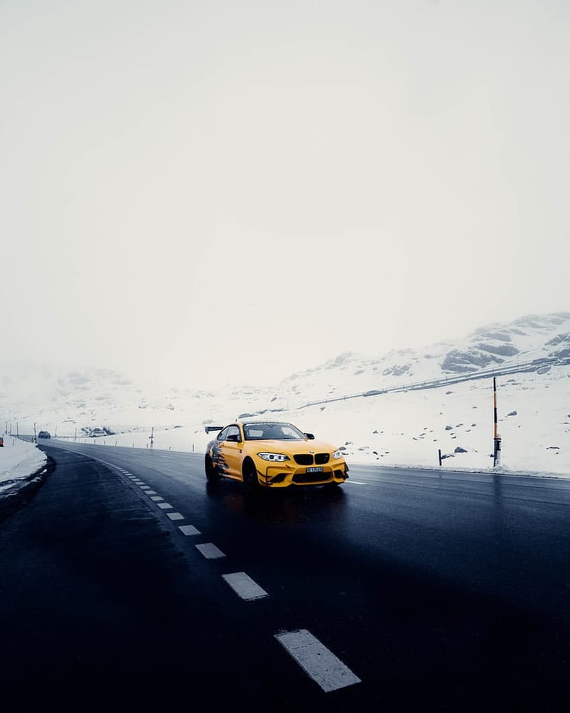 Yellow BMW M2 coupe with a rear spoiler driving on a wet mountain road between snow-covered hills under grey clouds.