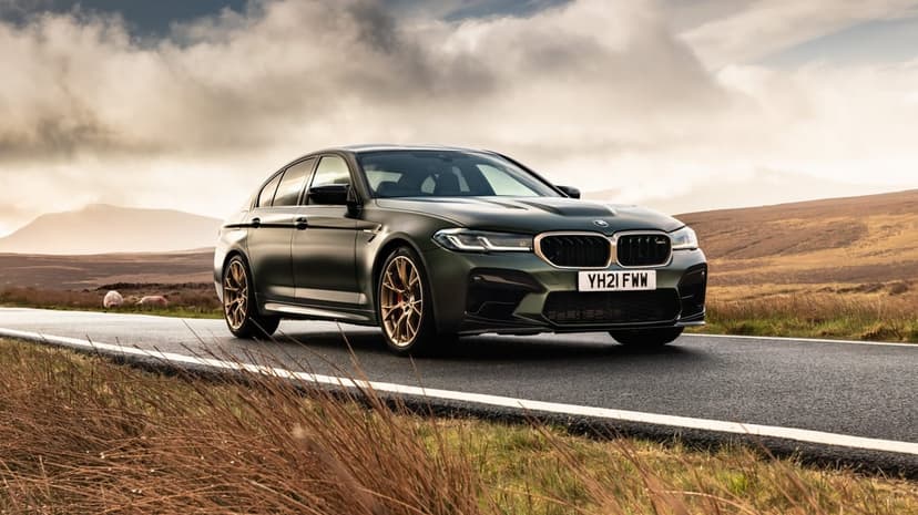 Matte green BMW M5 CS with bronze wheels on a rural road under a dramatic cloudy sky during golden hour.