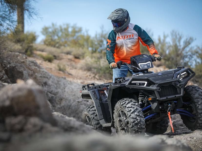 Black Polaris ATV climbing a rocky hill with a rider in an orange jersey under a clear blue sky.