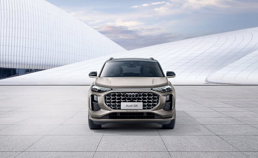 Front view of a beige Audi Q6 parked on a tiled plaza before sweeping white futuristic buildings under a soft sky.