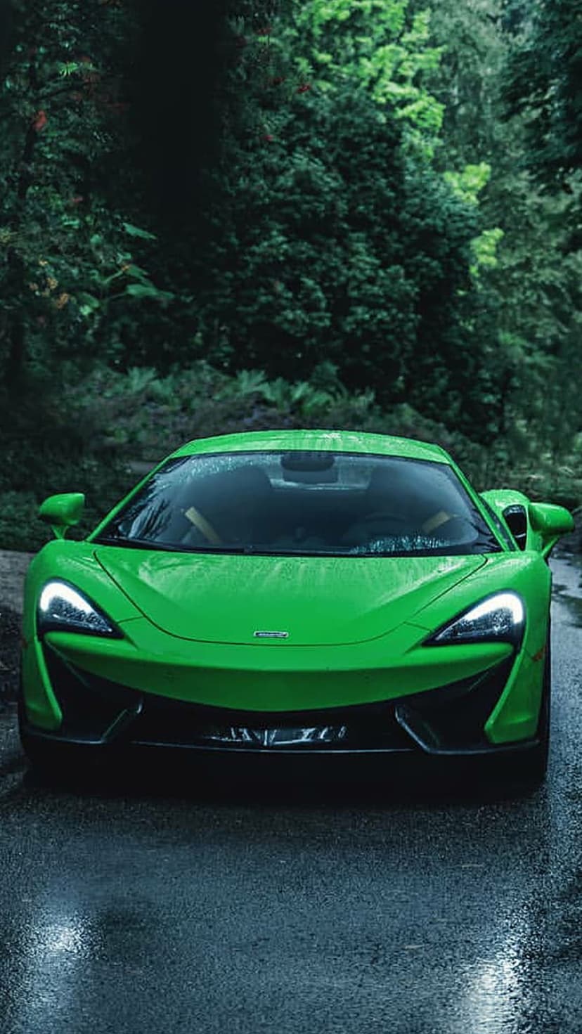 Green McLaren supercar with glowing headlights parked on a wet forest road under dark trees during a rain shower.