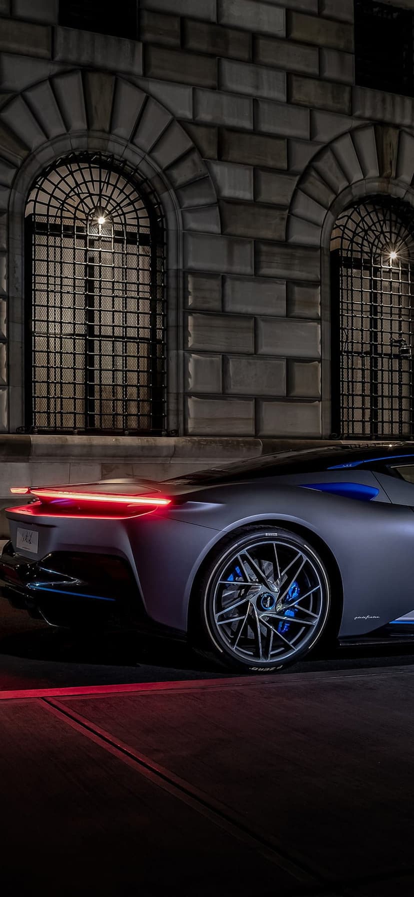 Gray electric hypercar with glowing red taillights parked in front of a stone building with arched windows.