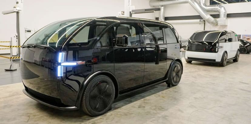 Black Canoo electric van with T-shaped LED lights in a bright industrial studio with a white van behind.