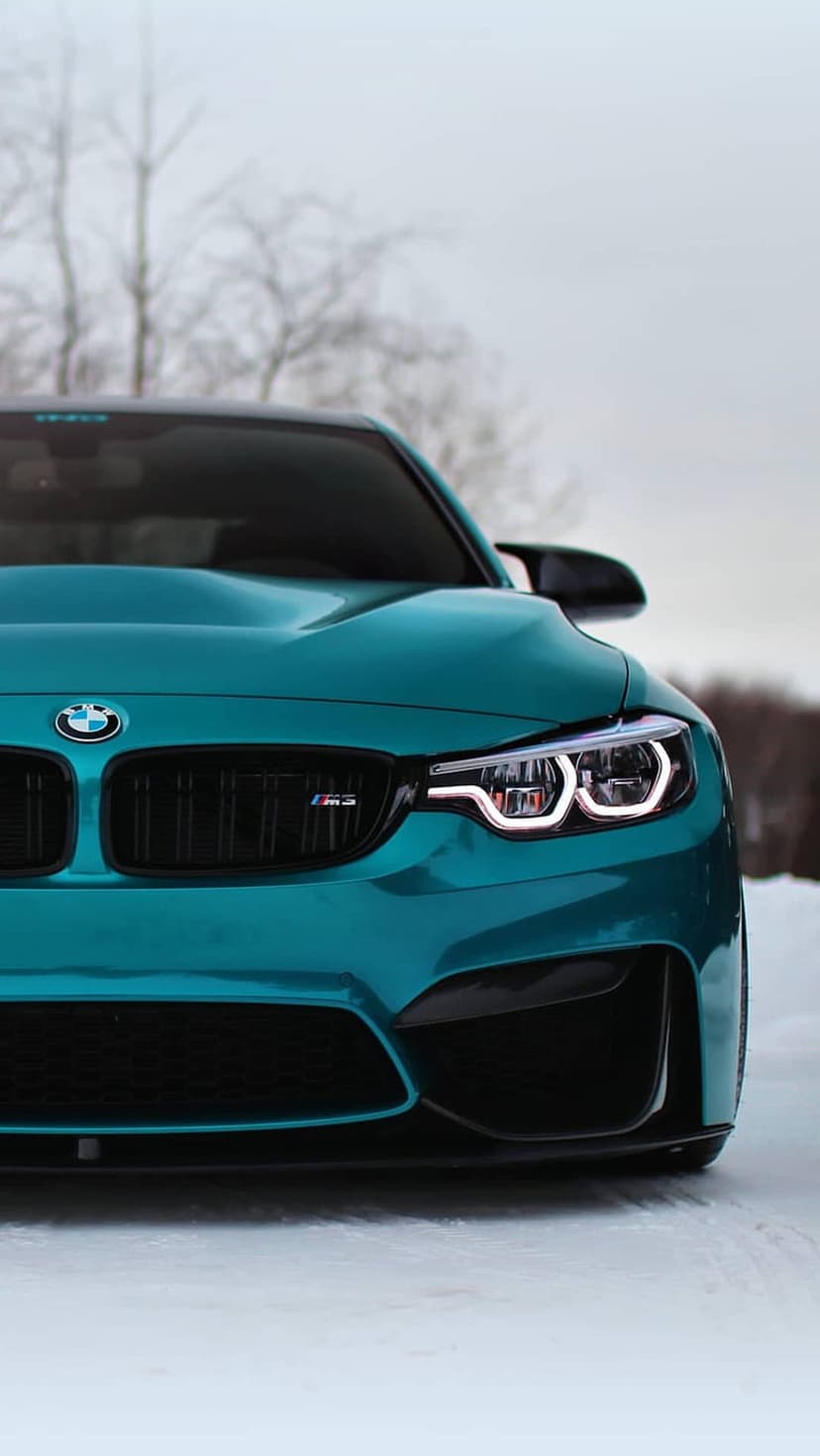 Teal BMW M4 with glowing LED headlights and a signature kidney grille parked on a fresh layer of winter snow.