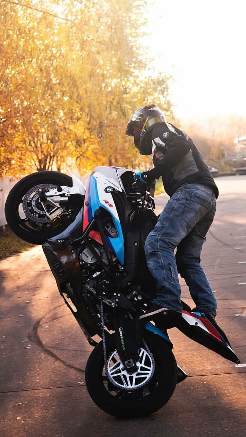 Motorcyclist in racing gear performs a wheelie on a white and blue BMW bike past vibrant orange autumn trees.