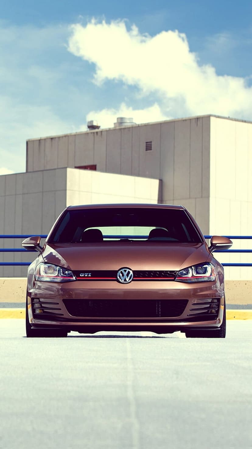Bronze Volkswagen Golf GTI with LED headlights parked in front of a beige modern building under a blue sky.