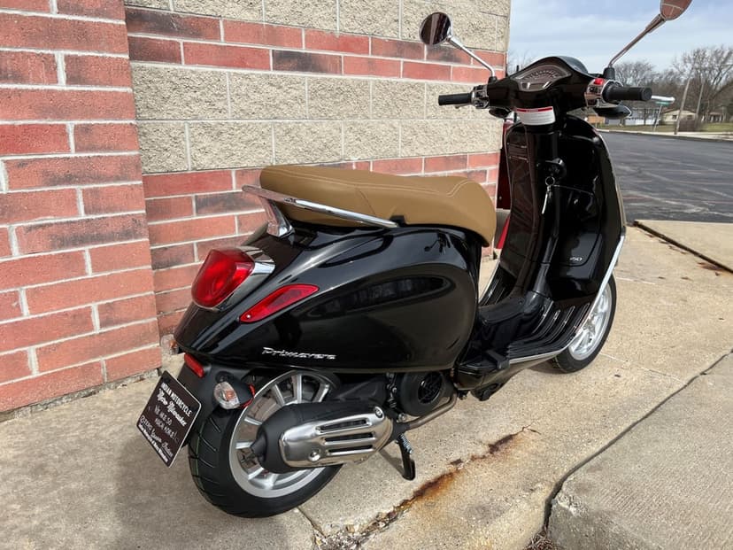 Black Vespa Primavera scooter with tan seat parked on concrete beside a red brick wall under soft daylight.