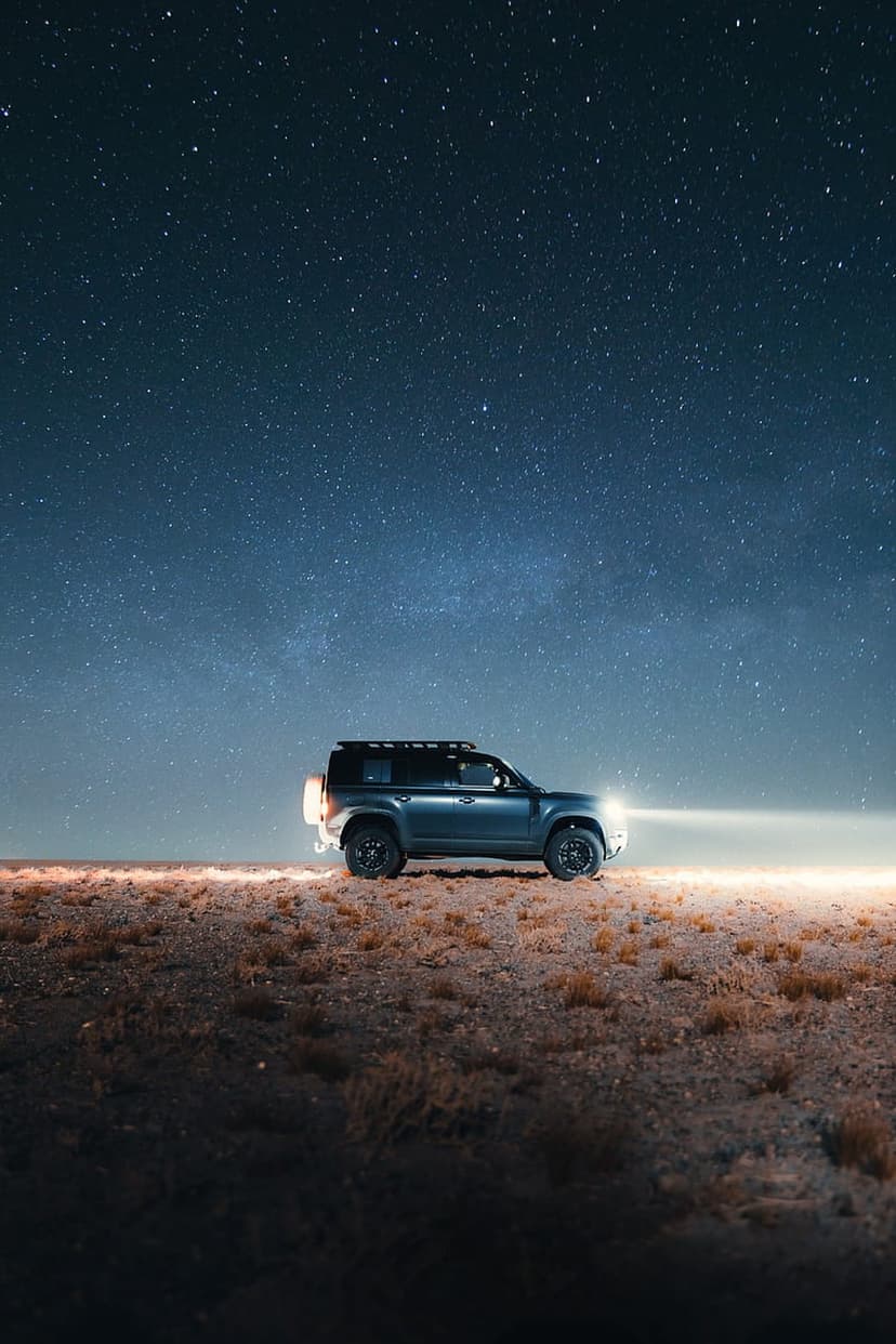 Land Rover Defender SUV parked on grassy terrain under a vivid Milky Way galaxy and starry night sky at midnight.