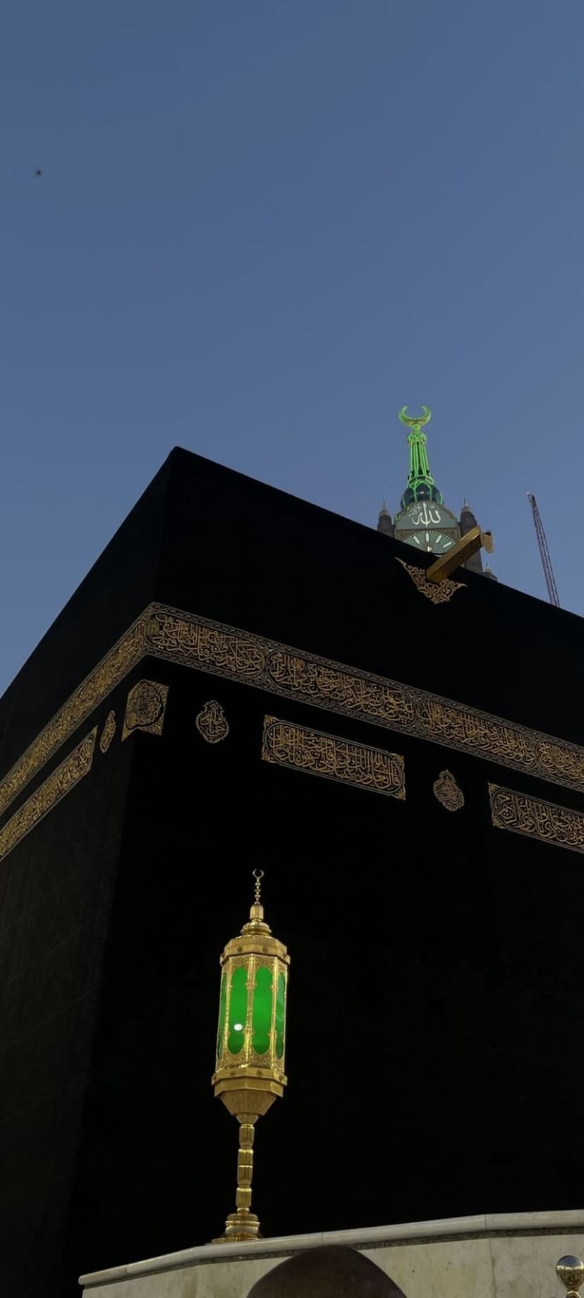 The Kaaba with gold calligraphy stands behind a green glass lantern under a crescent moon spire against a blue sky.