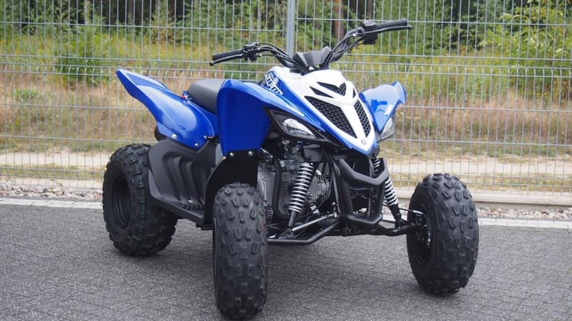 Blue and white Yamaha Raptor ATV parked on gray asphalt with off-road tires and a fence background.