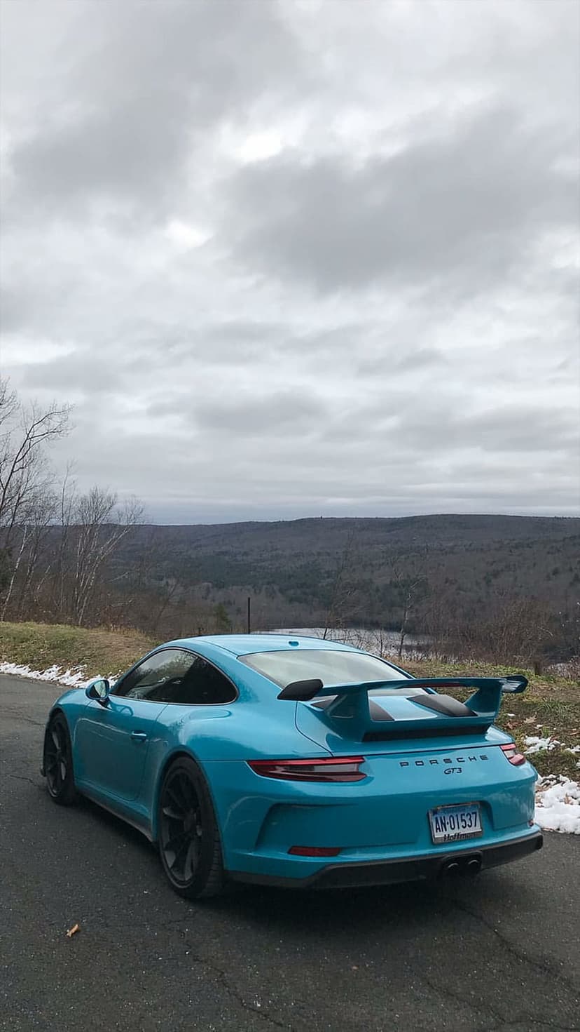 Turquoise Porsche 911 GT3 rear wing and black wheels on a snowy mountain road with a cloudy forest background.