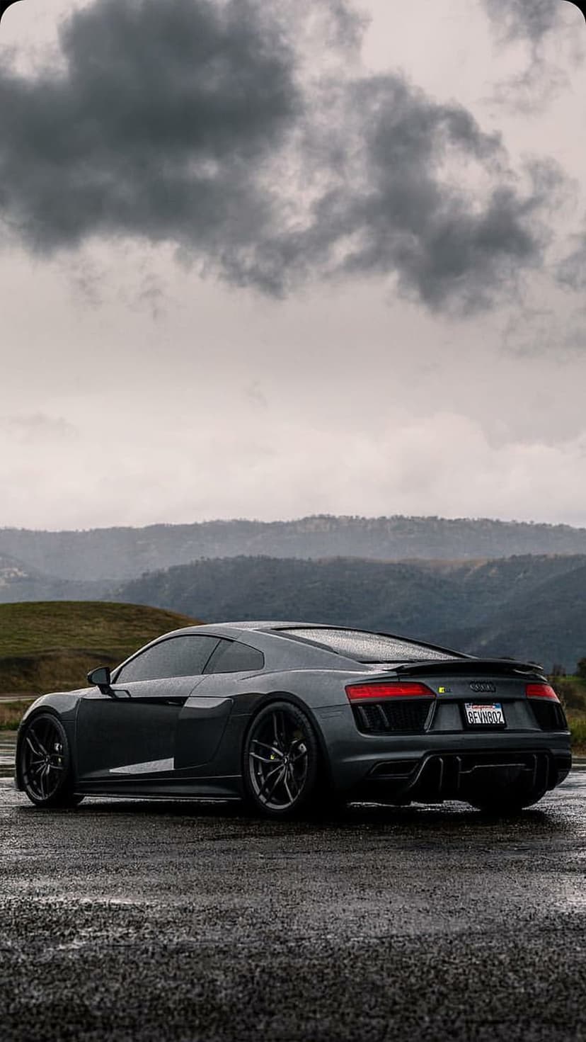Audi R8 supercar parked on a wet asphalt road under a dramatic, cloudy sky with distant hills and reflections.