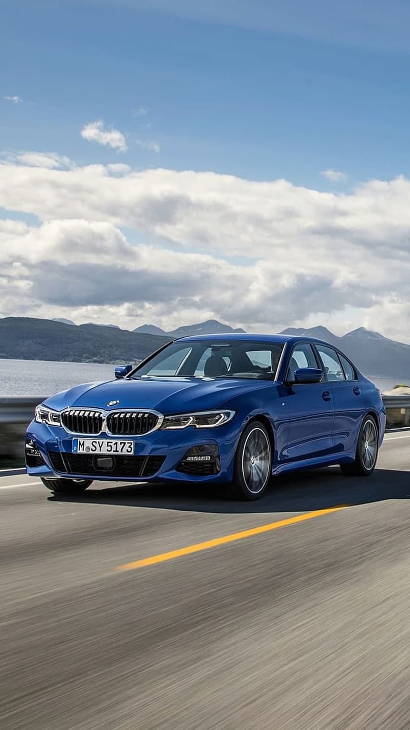 Blue BMW 3 Series sedan drives along a coastal highway with jagged mountains and a calm bay in the background.