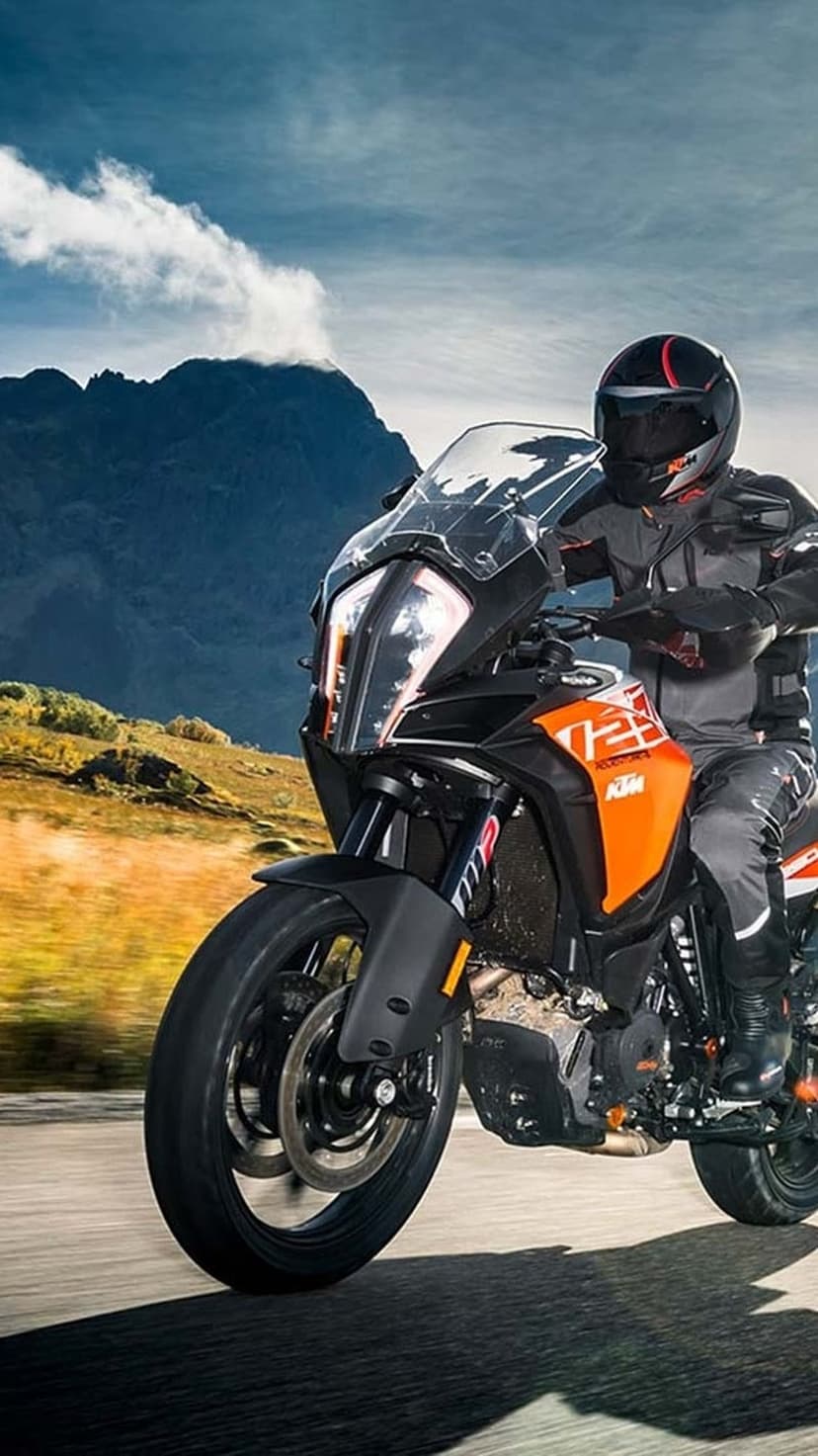 KTM rider on an orange adventure bike leans into a paved mountain road curve under a cloudy blue sky.