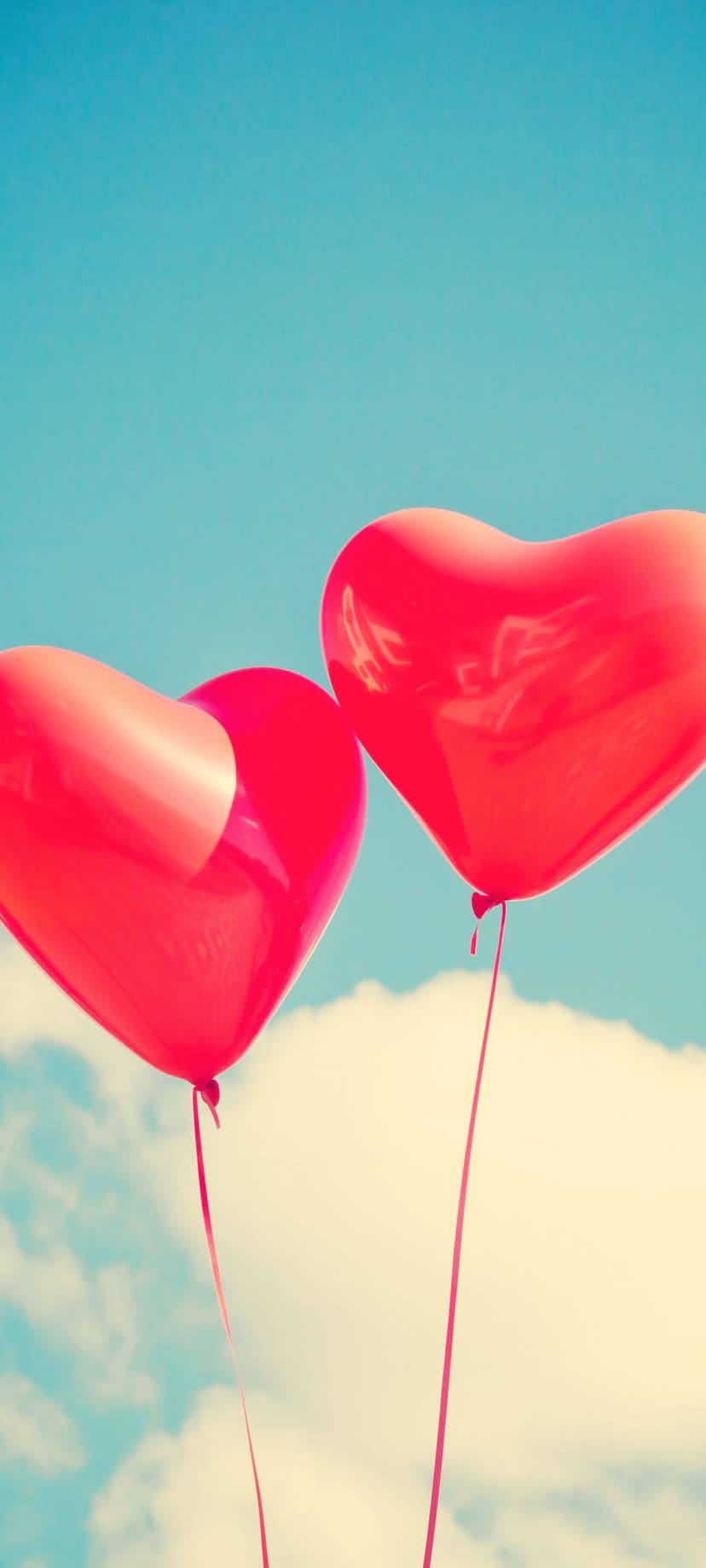 Two red heart-shaped balloons with thin ribbons float toward white clouds in a bright blue summer sky.