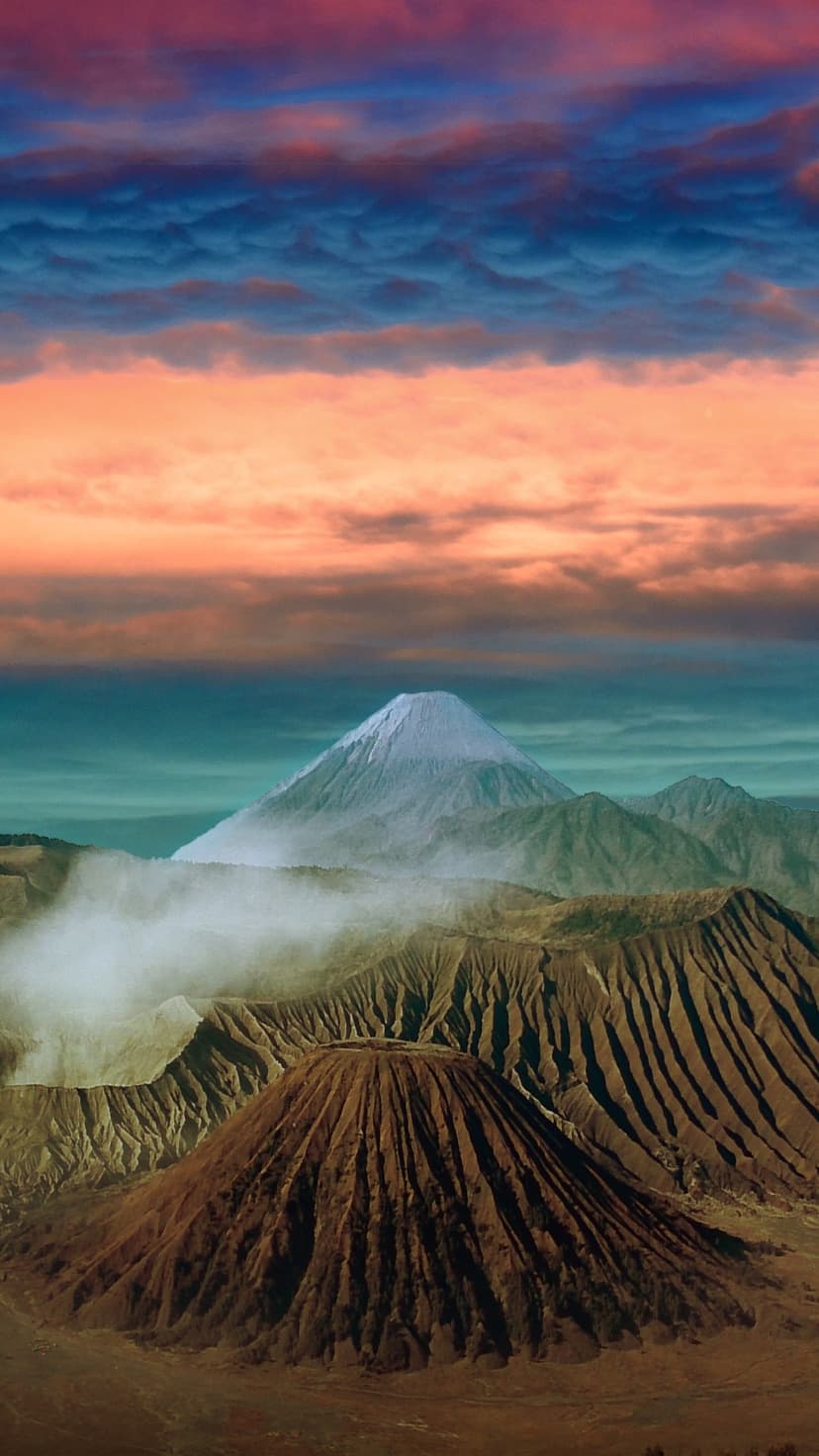 Snow-capped volcanic peak towering over mist-filled craters under a vibrant orange and blue sunrise sky.