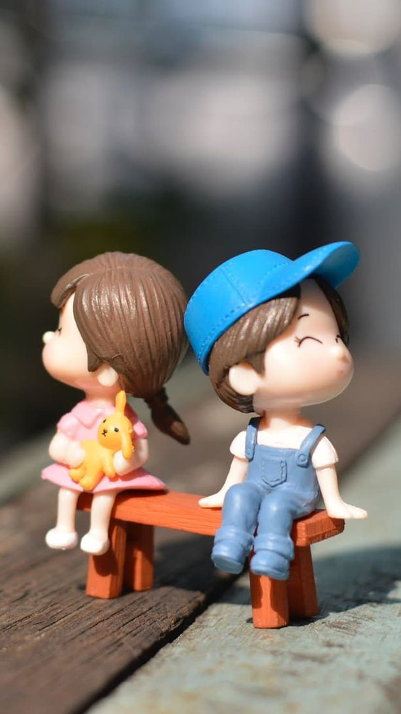 Miniature boy and girl figurines sit together on a small wooden bench with a yellow bunny toy and blue cap.